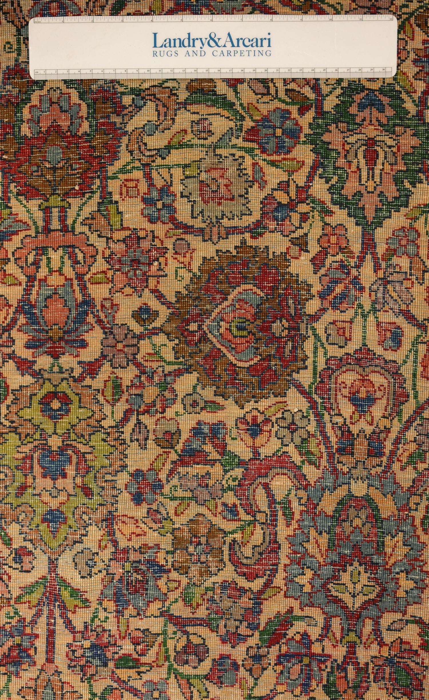 Close-up detail of a traditional Kerman rug's intricate floral pattern in muted reds, blues, and greens on a beige background.
