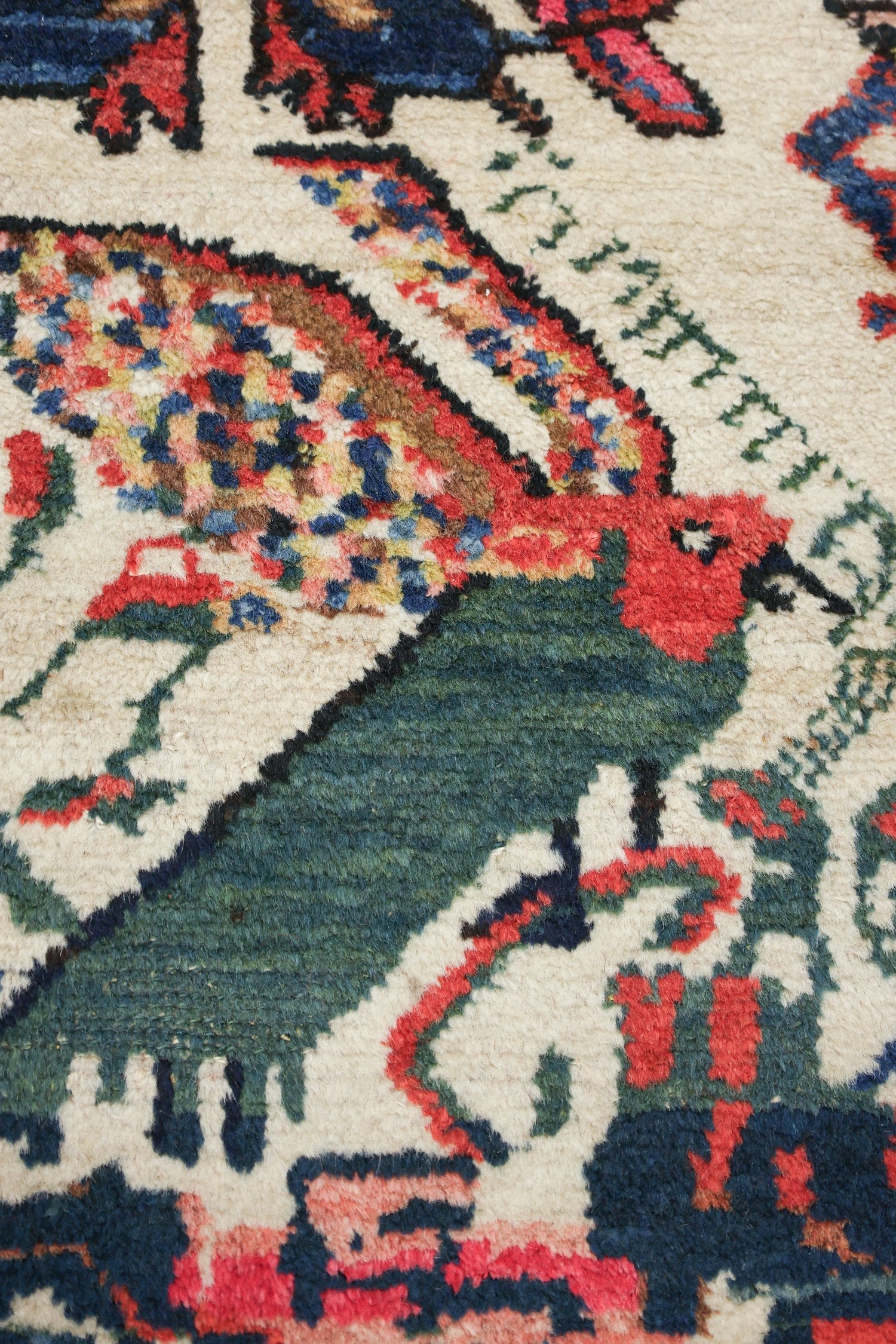 Close-up of antique rug detail: handwoven bird motif in red, green, and blue on an off-white background.
