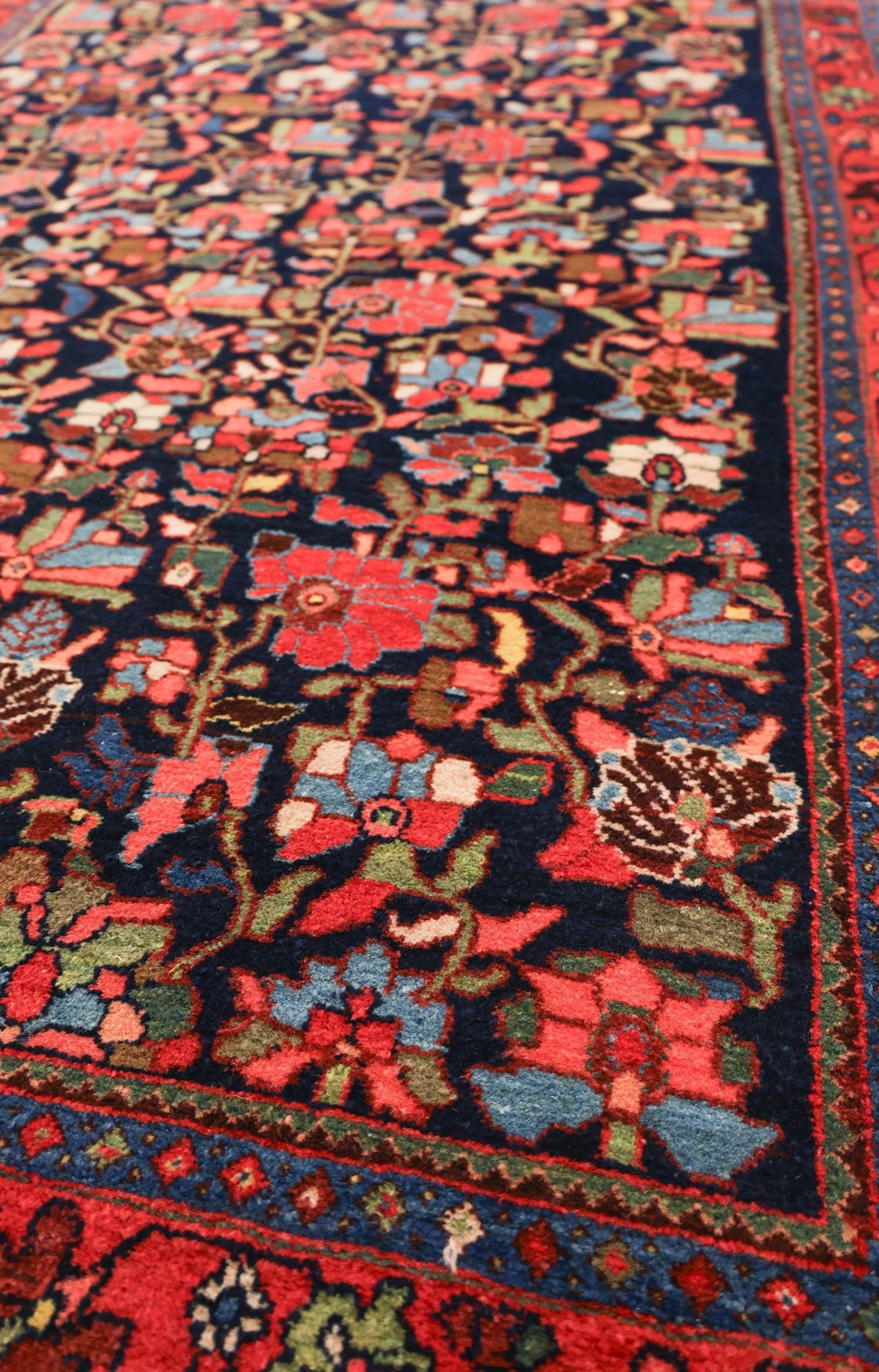 Close-up of a richly colored antique rug, showcasing intricate floral patterns in red, blue, and green on a dark navy background.
