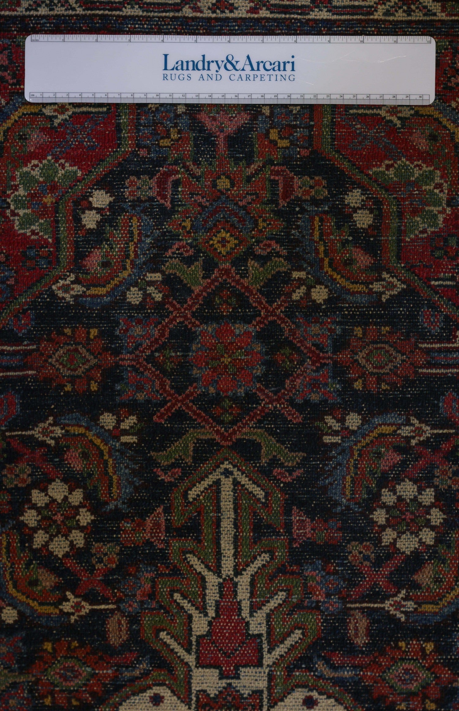 Detailed close-up of an antique rug's intricate, handwoven design featuring rich reds, blues, and greens on a dark background.
