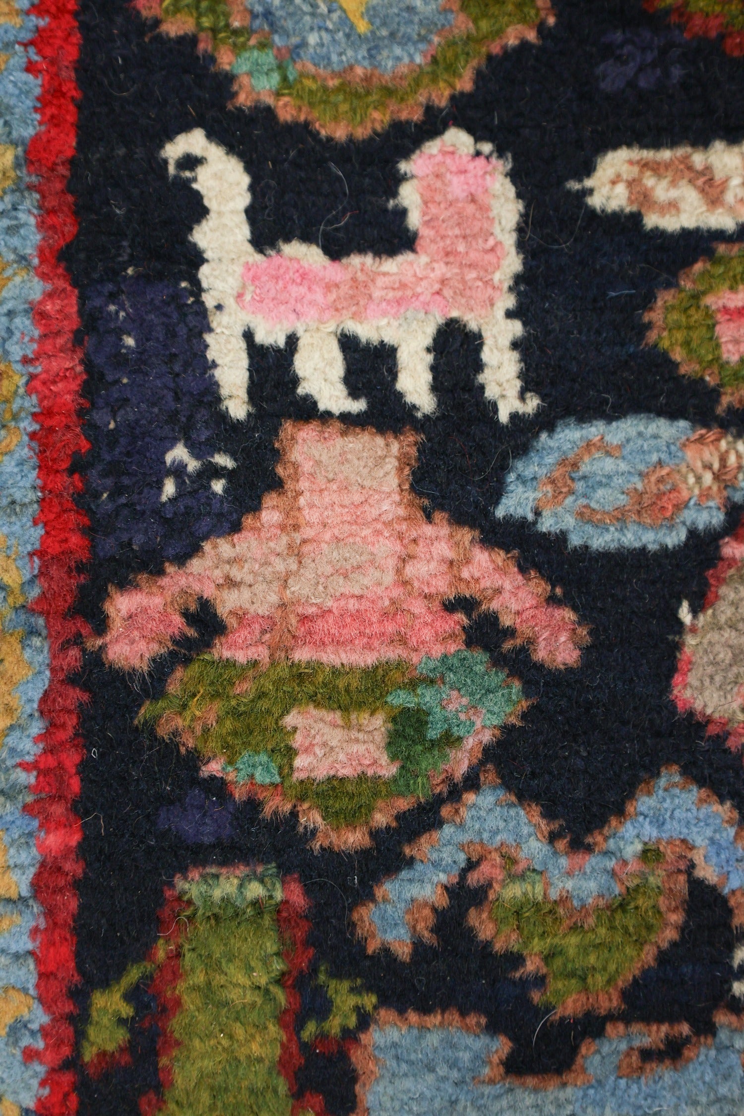 Close-up detail of a handwoven rug, showcasing a pink animal and multicolored floral patterns on a dark background.
