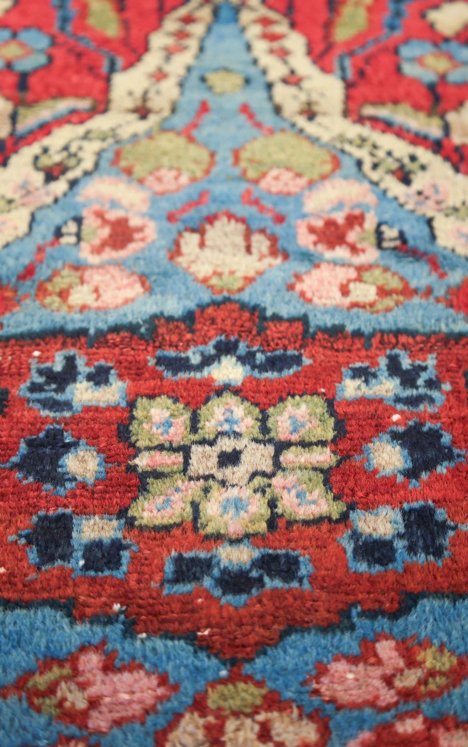 Close-up view of an antique rug's vibrant, handwoven floral pattern in red, blue, and beige.
