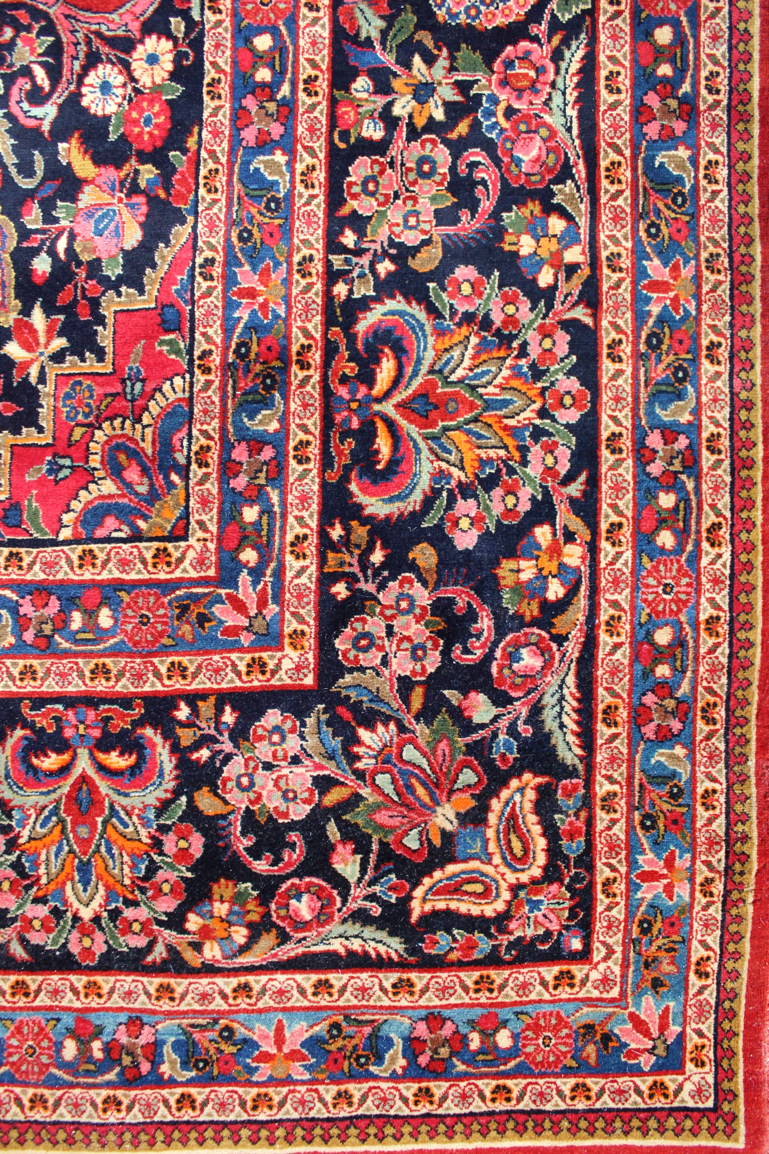 Detail of antique rug's border, showcasing vibrant floral and paisley patterns in rich reds, blues, and golds on a dark navy background.
