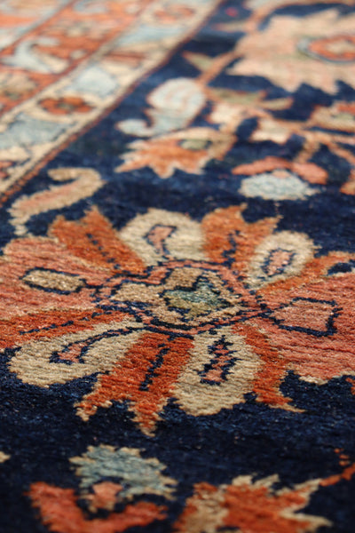 Close-up of antique rug detail: handwoven floral pattern in orange, beige, and navy blue.
