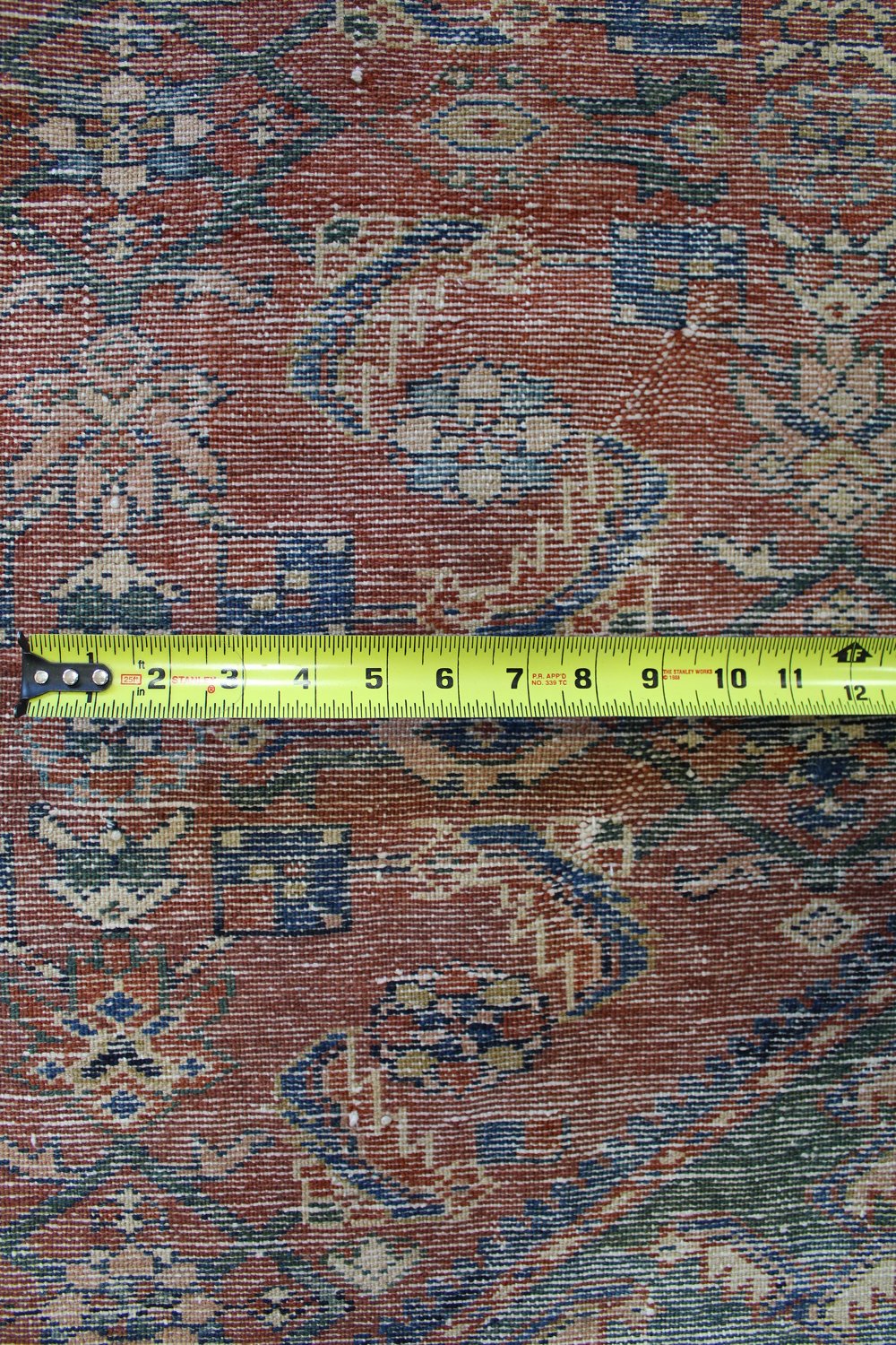 Close-up detail of antique handwoven rug, showing intricate pattern and measuring tape for scale.
